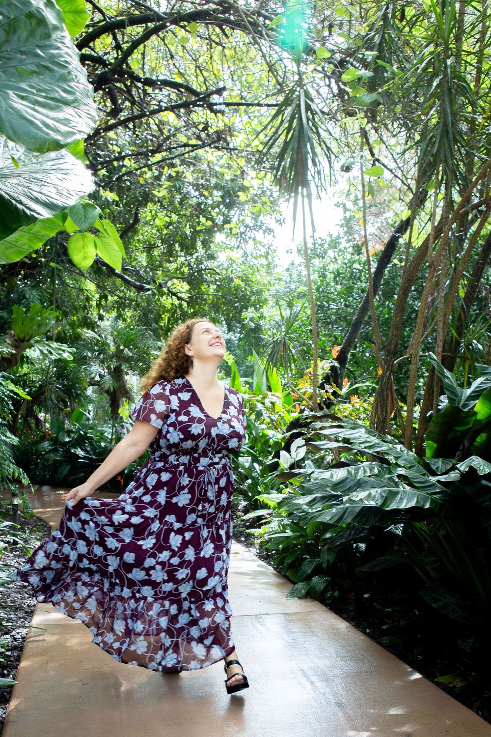 Woman in floral maxi dress walking joyfully through lush tropical garden path surrounded by green foliage.