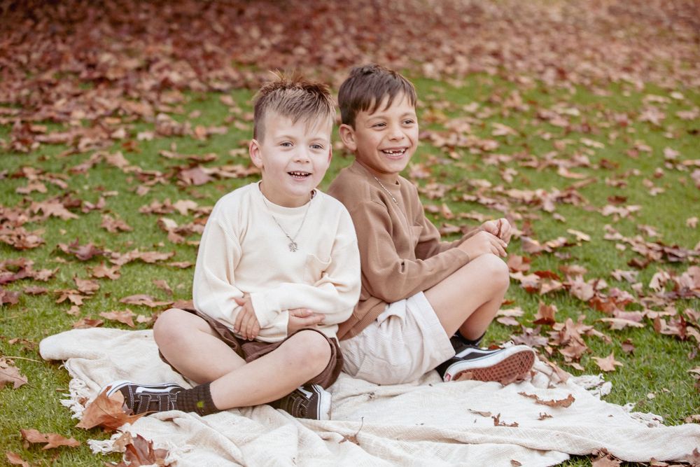 Two boys in neutral tones sit together on a blanket surrounded by autumn leaves on green grass.