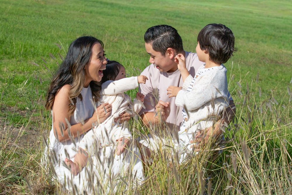 Family of four sitting in a green field, parents interacting with two young children in matching white outfits.