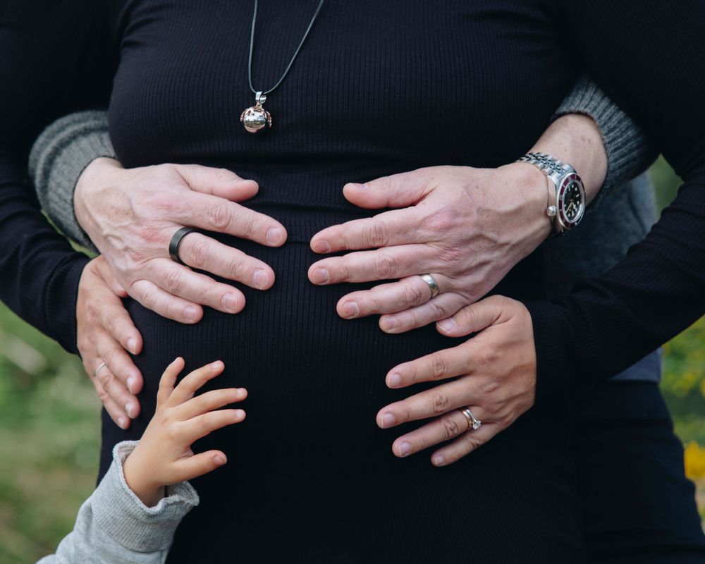 Close-up of hands touching a pregnant belly in black clothing against a blurred outdoor background.