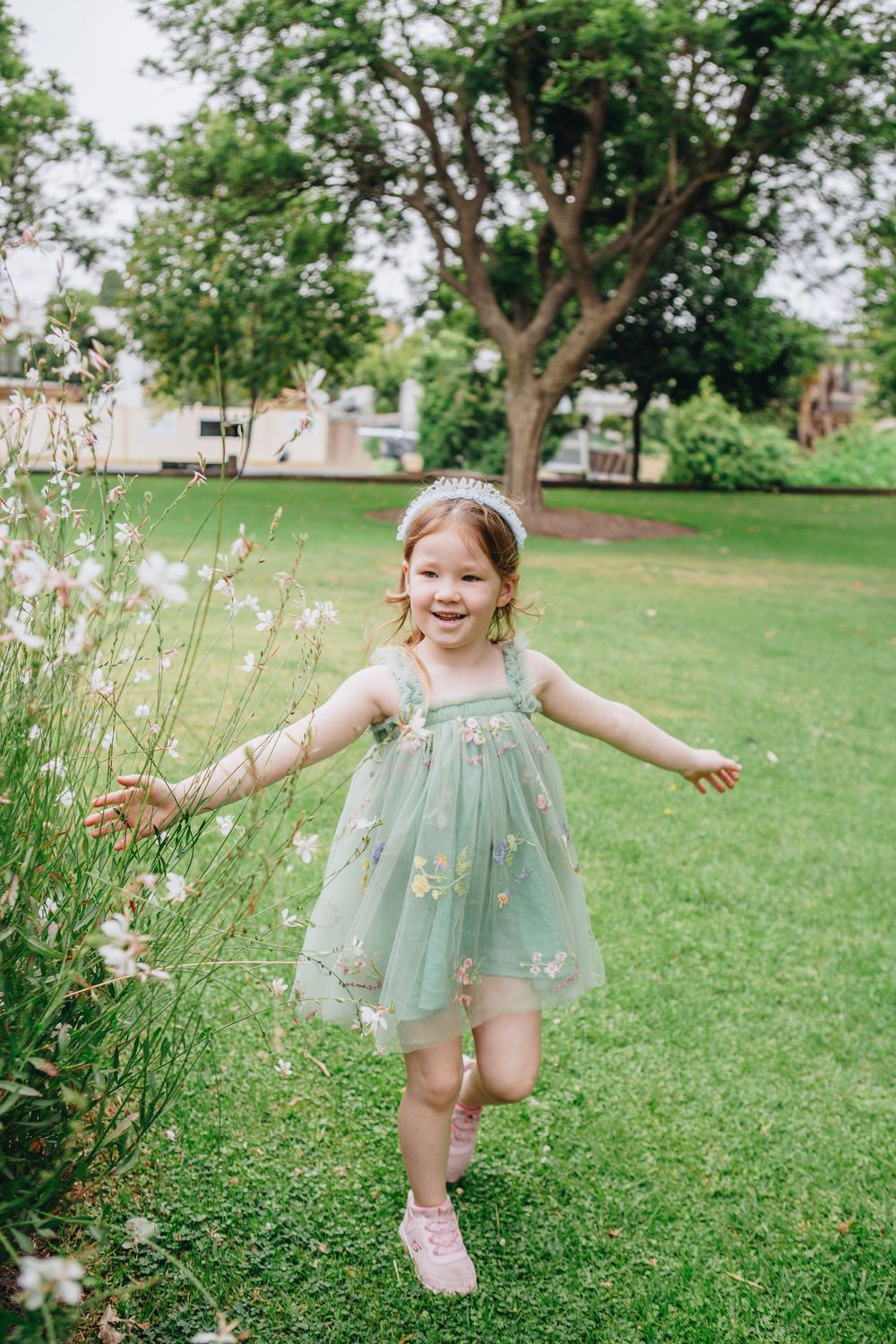 Happy young girl in green floral tulle dress runs through a garden wearing a silver tiara.