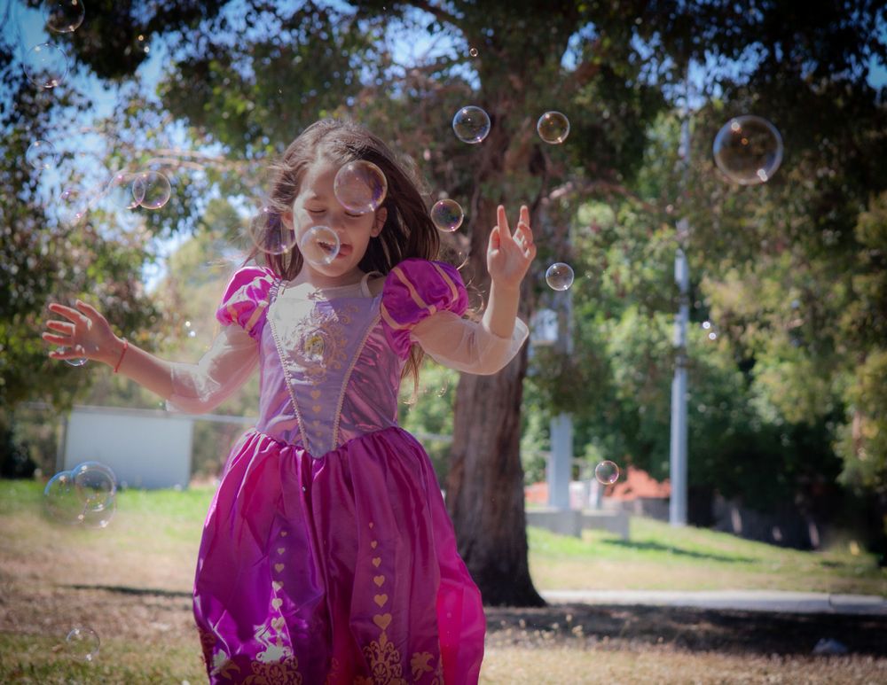 Young child in princess dress jumping and playing with bubbles in a backyard setting.