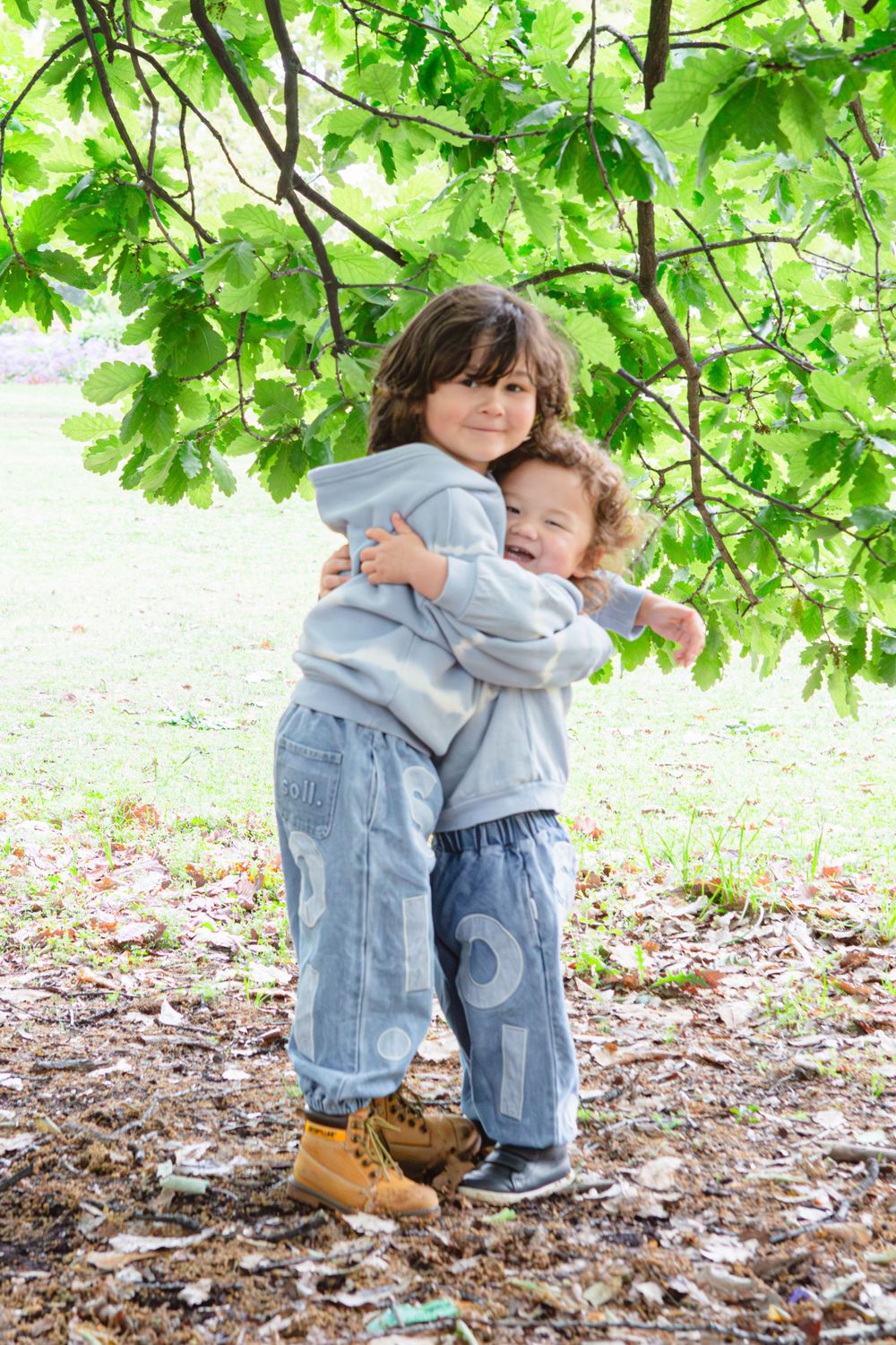Two young boys in matching denim outfits share a warm hug under a leafy tree in a park.