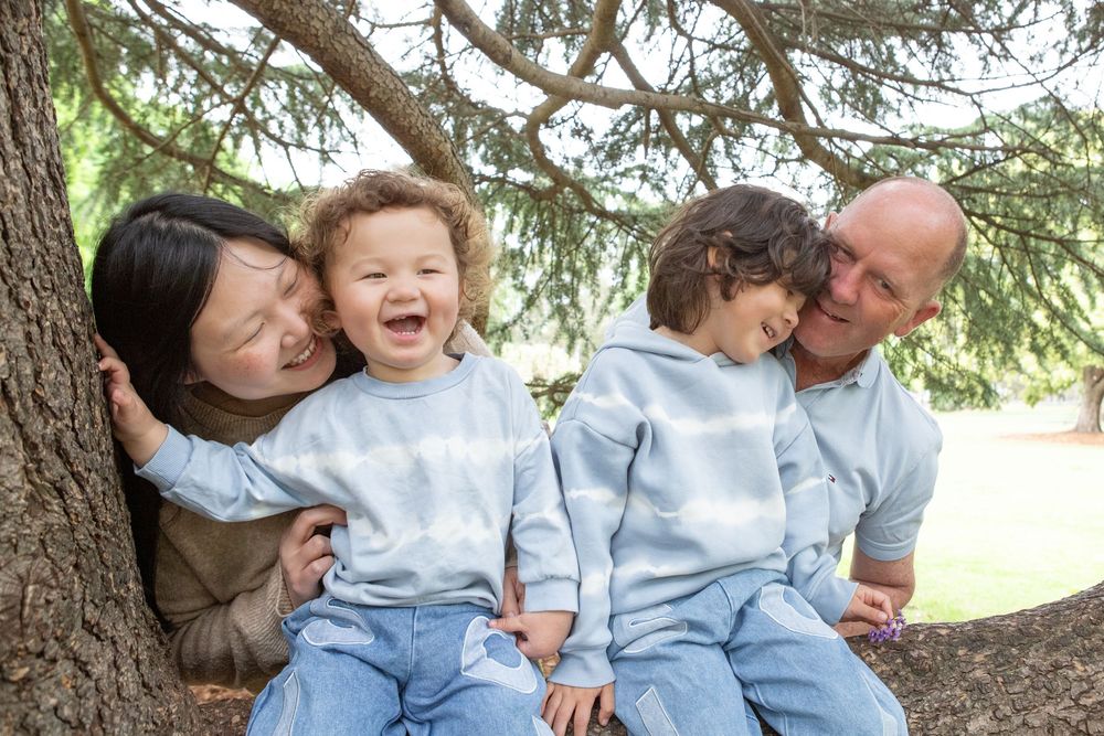 Family laughing and playing together while sitting under a large evergreen tree on a sunny day.