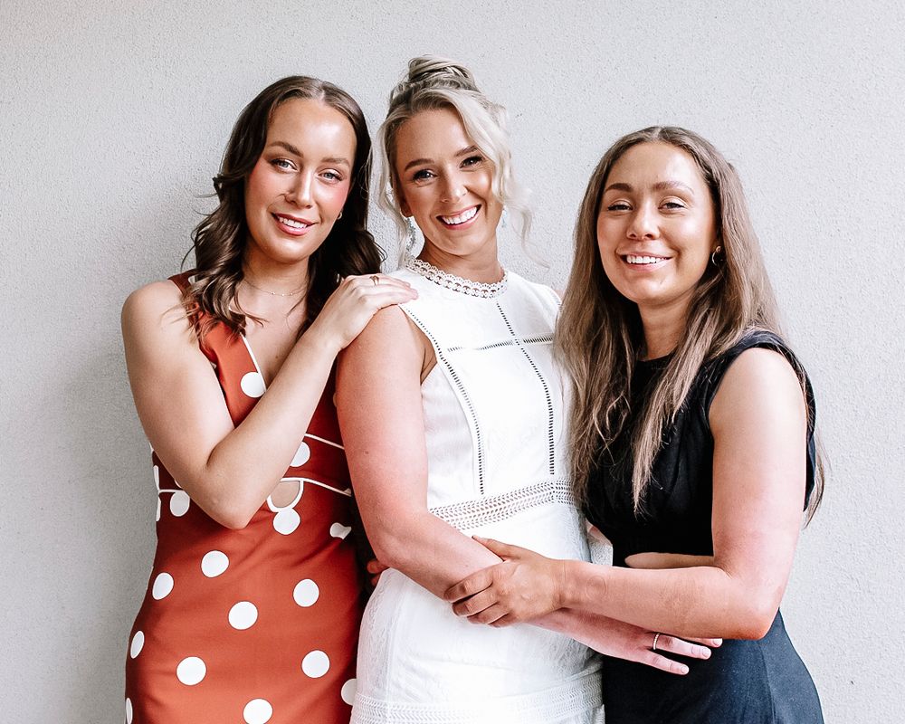Three women in casual attire smiling and posing together against a white background.