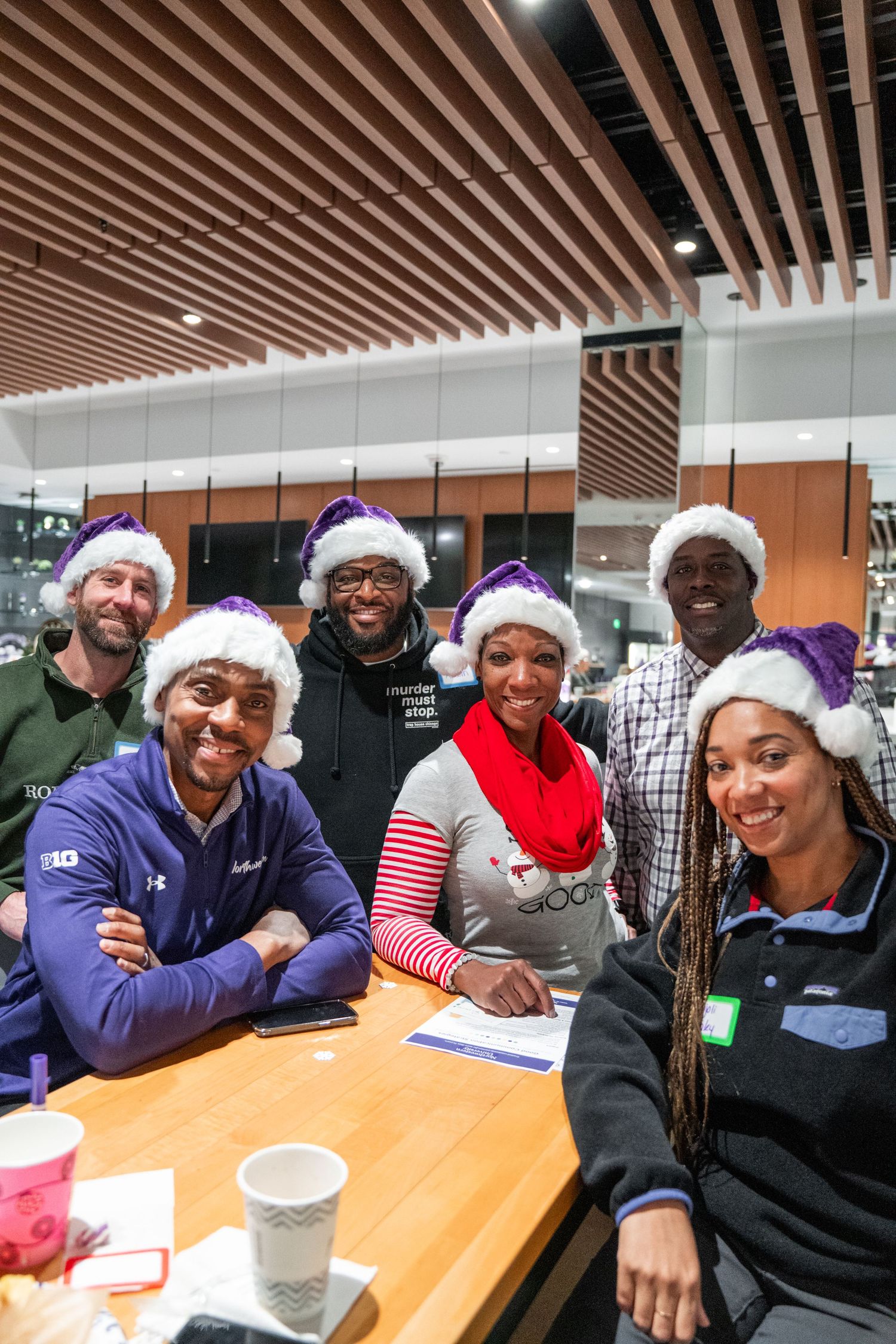 Group wearing purple Santa hats gathered around table for holiday celebration.