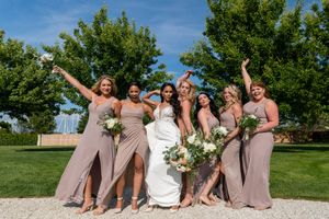 Bride with bridesmaids in taupe dresses celebrating outdoors on a sunny day with bouquets.