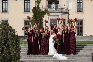 Bridesmaids in burgundy dresses celebrate with bride on grand steps of elegant venue.