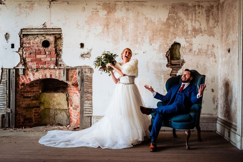 Bride holds bouquet as groom reacts with joy in rustic venue with exposed brick fireplace.