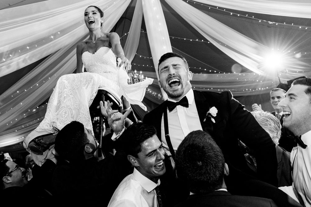 Bride and groom lifted by joyful guests during energetic wedding reception celebration.