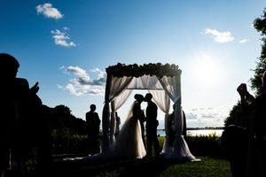 Silhouette of couple kissing under floral arch at outdoor wedding ceremony at sunset.
