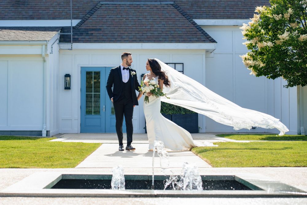 Bride's veil flows in wind as couple poses by fountain outside white wedding venue.