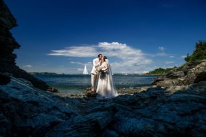Wedding couple kissing on rocky coastal shoreline with sailboat and blue sky backdrop.