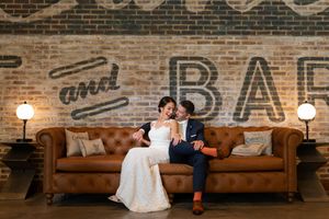 Bride and groom sitting on leather couch in front of rustic brick wall with bar signage.