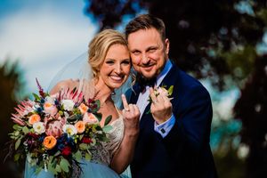Smiling bride and groom showing wedding rings with colorful bridal bouquet outdoors.