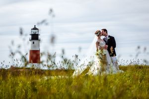 Bride and groom kissing in tall grass with iconic red and white lighthouse in background.