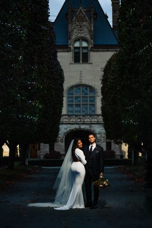 Bride and groom posing before a grand Gothic castle surrounded by tall trees at dusk.