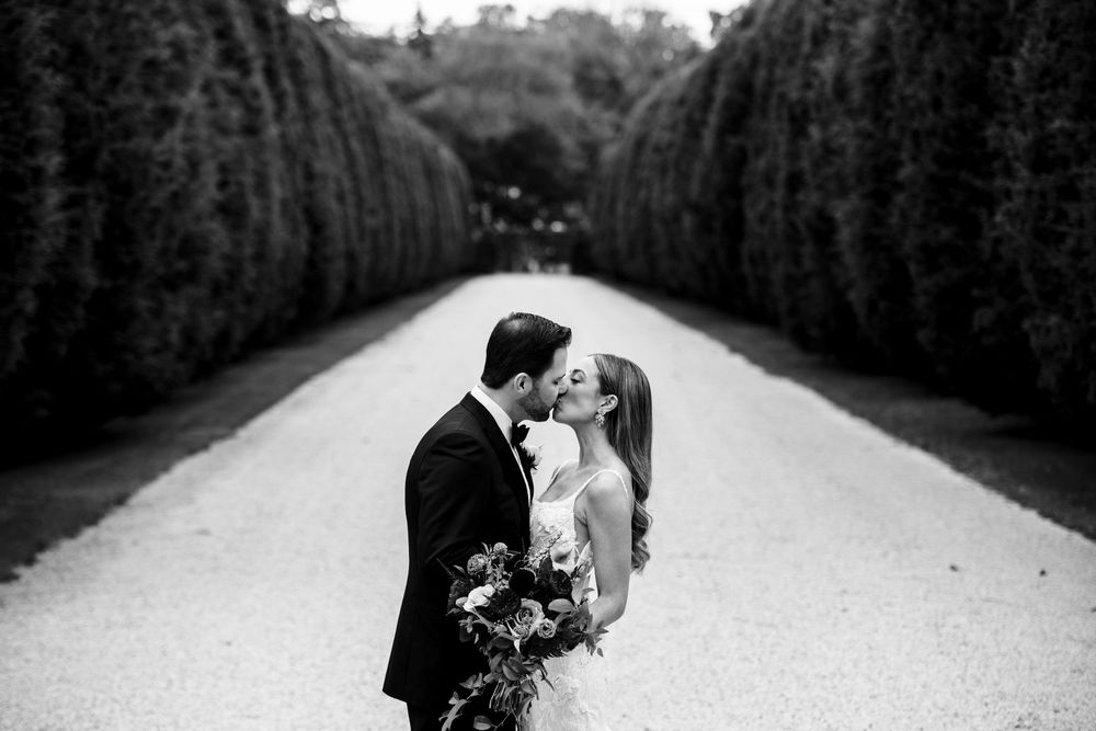 Black and white photo of bride and groom kissing on a grand tree-lined garden pathway.