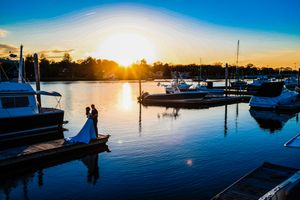 Bride and groom silhouetted on marina dock at golden sunset with sailboats reflected in calm water.