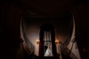 Elegant bride and groom silhouetted on grand curved staircase in dimly lit ornate wedding venue.