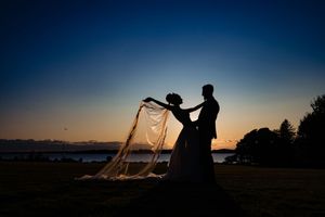 Romantic silhouette of bride and groom at twilight, wedding veil flowing in the breeze by the water.