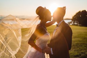 Bride and groom share a kiss bathed in golden sunset light, veil billowing dramatically in the wind.