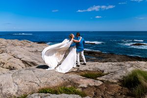 Bride in flowing white gown and groom dance on rocky coastal cliffs overlooking the blue ocean.