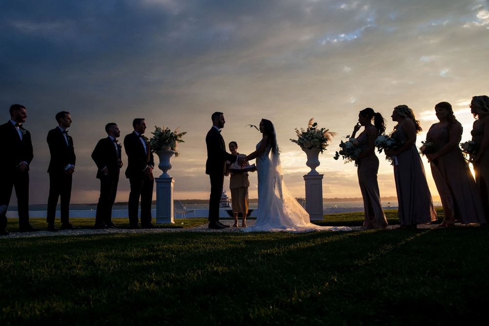 Wedding ceremony silhouette at sunset with bridal party lined up outdoors overlooking the water.