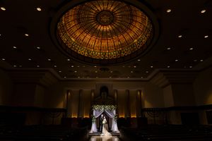 Bride and groom stand under a stunning stained glass dome during an elegant indoor wedding ceremony.