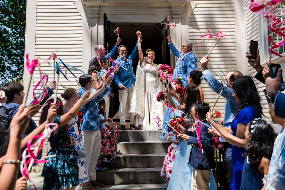 Joyful newlyweds celebrate with guests waving colorful ribbons outside a white chapel on a sunny day.