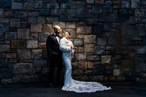 Bride and groom share a romantic moment against a dramatic stone wall in wedding attire.