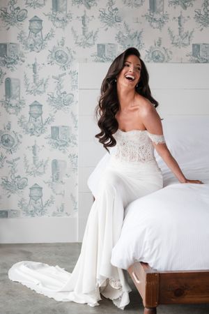 Radiant brunette bride in an off-shoulder lace wedding gown laughs joyfully in a decorated room.