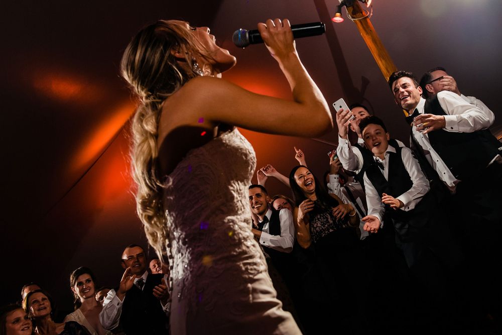 Bride passionately sings into a microphone while guests cheer and celebrate at a lively wedding reception.
