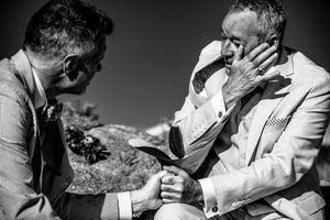 Emotional black and white wedding photo of groom holding hands with tearful elderly man outdoors.