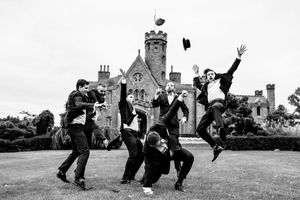 Groomsmen joyfully jumping and tossing hats in front of a castle venue in black and white.