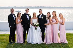 Wedding party in pink bridesmaid dresses and black tuxedos posing by waterfront at sunset.