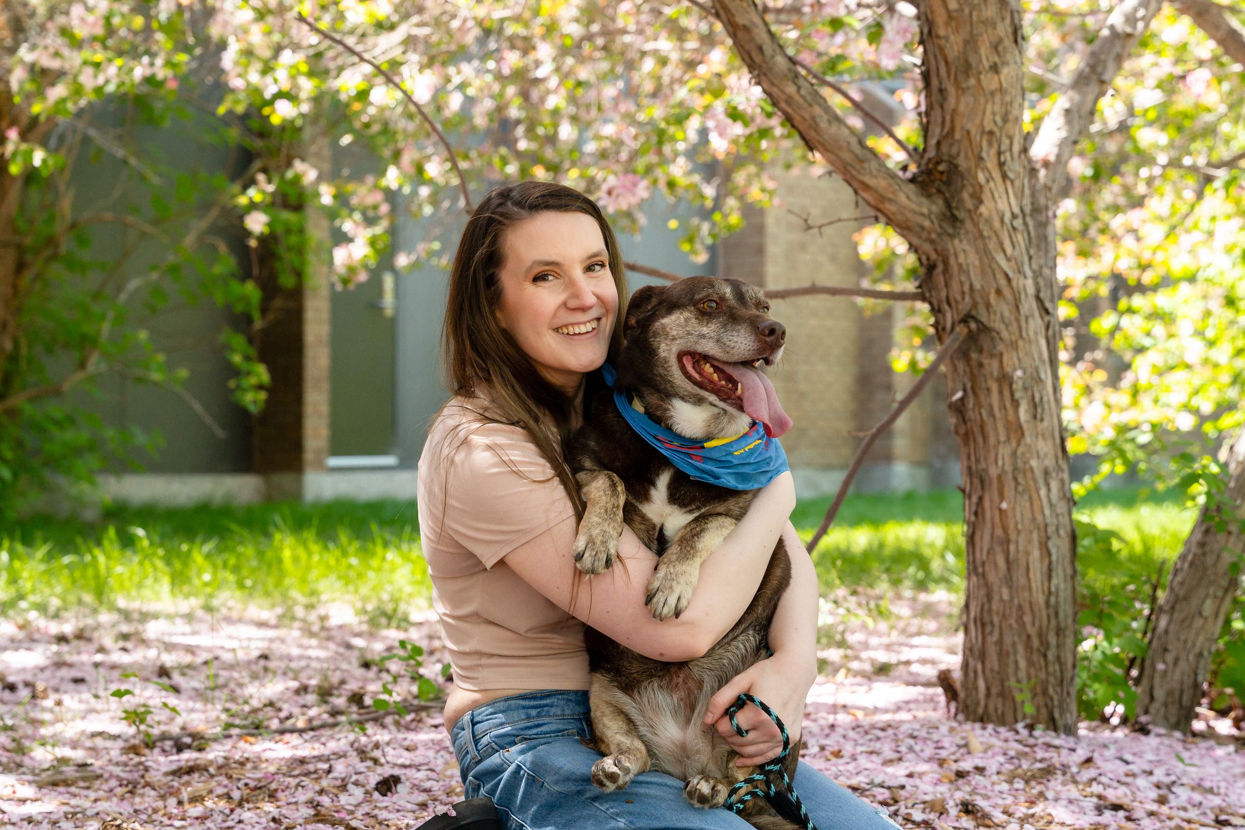 Person sitting outdoors cuddling with a happy dog wearing a blue bandana.