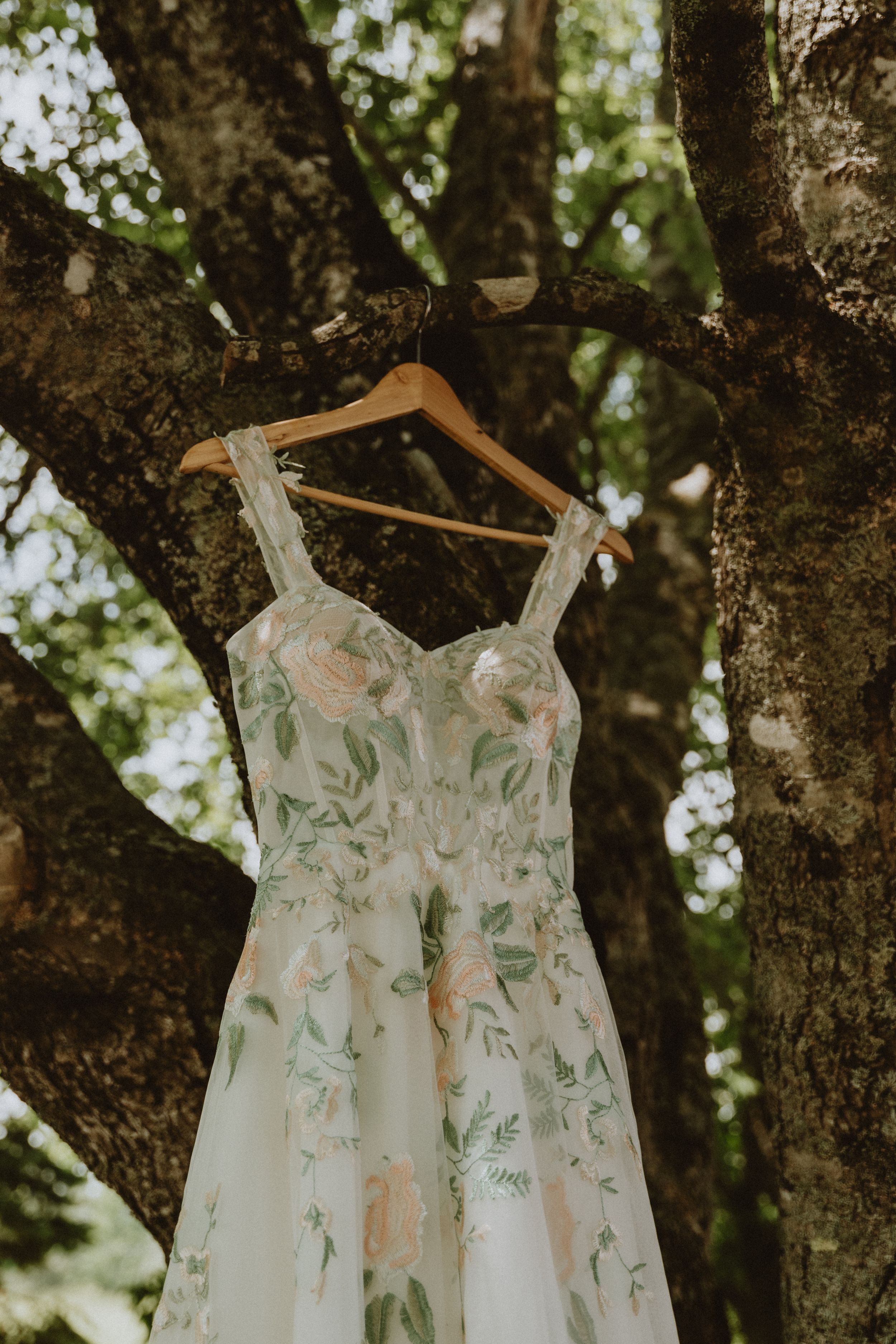 A delicate white wedding dress with floral embroidery hangs from a wooden hanger on a tree branch in natural lighting.