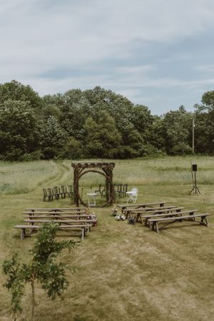 Rustic outdoor wedding ceremony setup with wooden benches and an arch in a grassy field surrounded by trees.