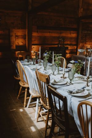 Rustic barn dining setup with wooden chairs and white tablecloth set for an elegant dinner under warm lighting.