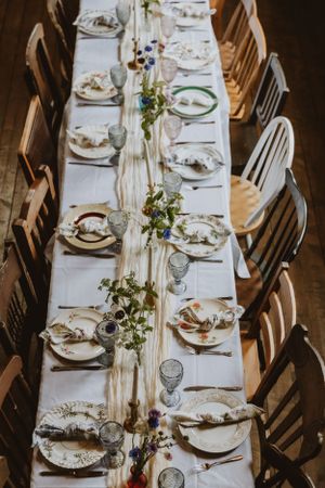 Elegant rustic wedding table setting with white tablecloth, wooden chairs and delicate greenery centerpiece runner.