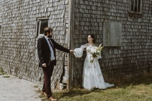 A couple in wedding attire poses against a rustic wood-shingled building wall in a vintage inspired setting.