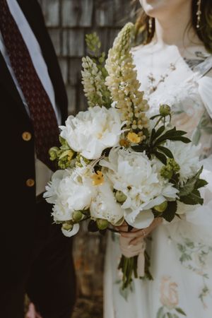 An elegant white and yellow wedding bouquet featuring peonies and delphinium flowers against formal attire.