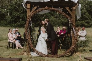 A rustic outdoor wedding ceremony under a large circular wooden arch decorated with twigs and branches in a field.