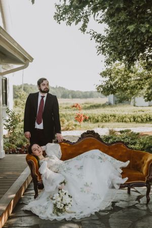 Couple in wedding attire posing on a vintage sofa on an outdoor porch.