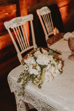 Vintage white chairs and lace tablecloth with elegant floral centerpiece featuring white hydrangeas and greenery.