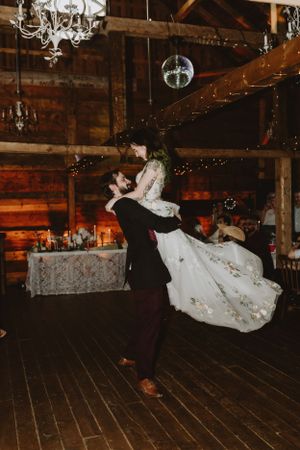 A couple shares their first dance in a rustic barn venue with twinkling lights and chandeliers overhead.