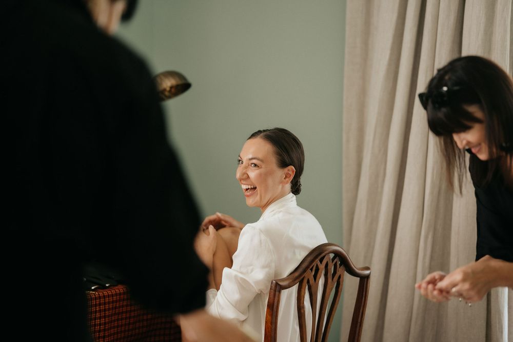 A person in a white shirt sits in a wooden chair smiling during what appears to be a photo session or event preparation.