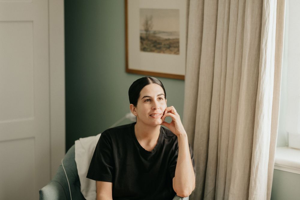 Person in black t-shirt sitting near window with thoughtful expression in room with green walls and framed artwork.