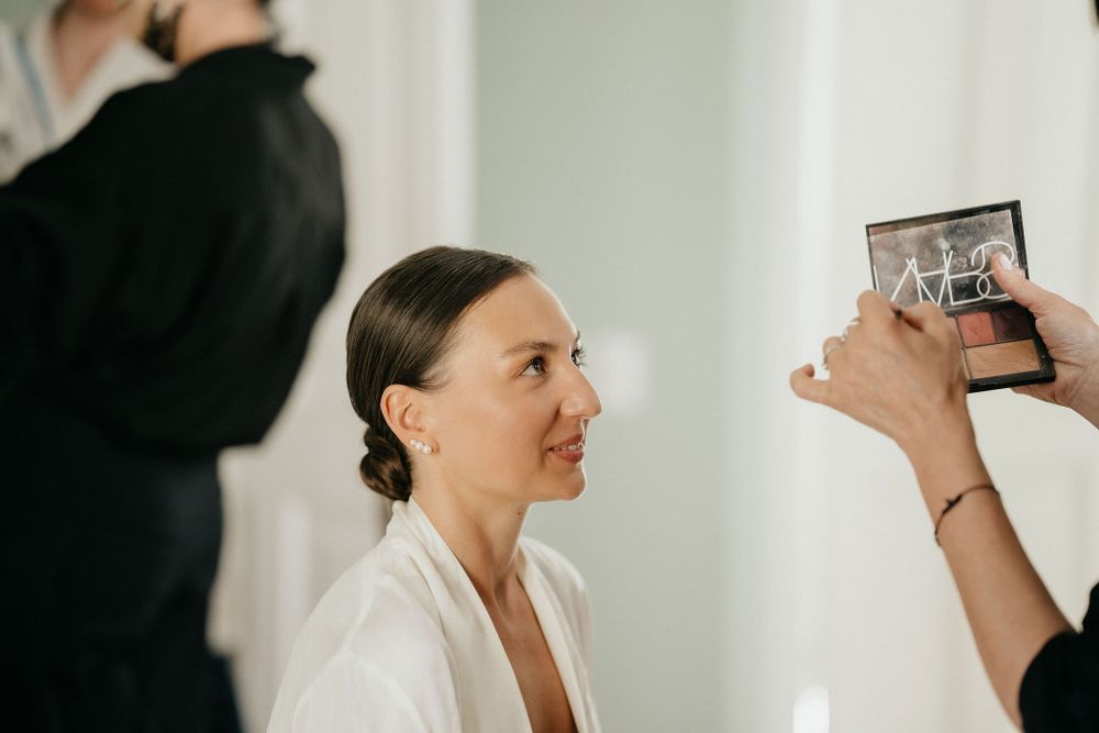Someone has their makeup done while looking at a phone being held up as a mirror during a preparation session.